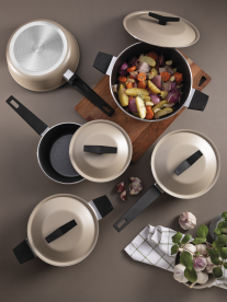 Top-down view of a beige cookware set with multiple pots and lids arranged on a table, one pot filled with roasted vegetables on a wooden board, with garlic and herbs nearby.