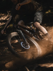 A person riding a mountain bike on a dirt trail in a forest, kicking up dust while turning.