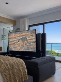 A modern bedroom with a large TV at the foot of the bed, ocean view through glass doors, and a bathroom visible in the background.