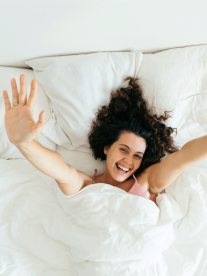 A woman with brown curly hair, lying in a comfortable bed from The Bed Centre with crisp white sheets.