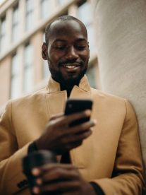 A cool black male wearing a stylish camel coat, searching on his cellphone and smiling while he stands against the wall of a building