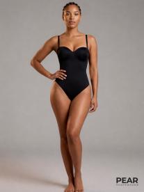 A woman stands barefoot in a studio wearing a black shapewear bodysuit against a light grey background, with the Pear Shapewear logo in the corner.
