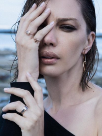 A brunette woman wearing Verus Fine Jewellery rings, and a black one-sided top stood on a beach.