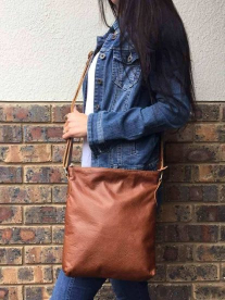 A lady holding a tan Mirelle genuine leather everyday messenger-handbag as she stands in front of a brick wall.