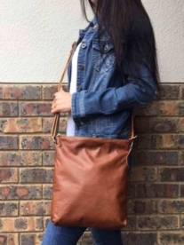 A lady holding a tan Mirelle genuine leather everyday messenger-handbag as she stands in front of a brick wall.
