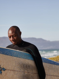 A black man with his surfboard at the beach on his way to surf on a white and blue surfboard